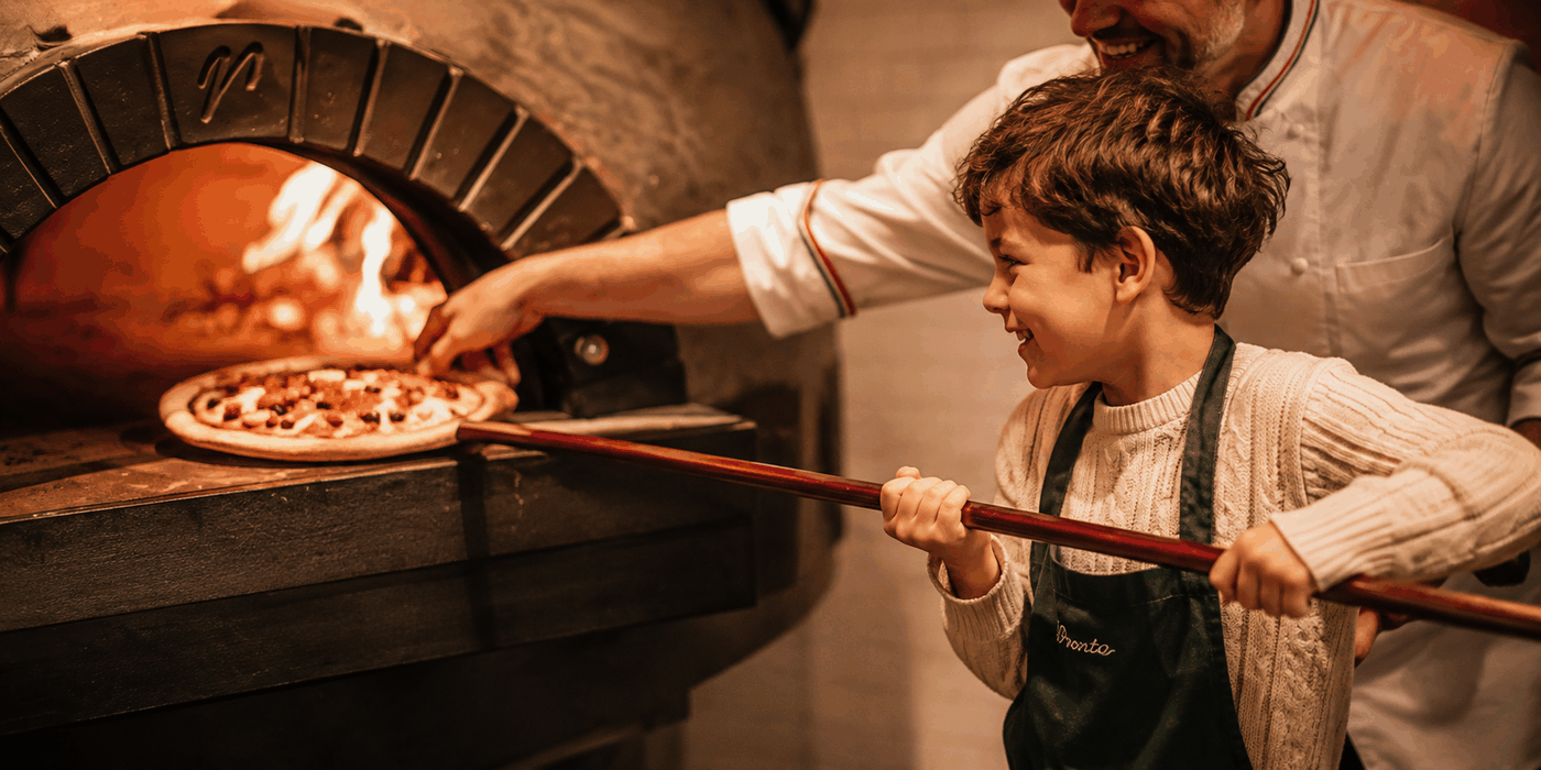 Jongen schuift met de pizzaschep een pizza in de houtgestookte steenoven, samen met de chef